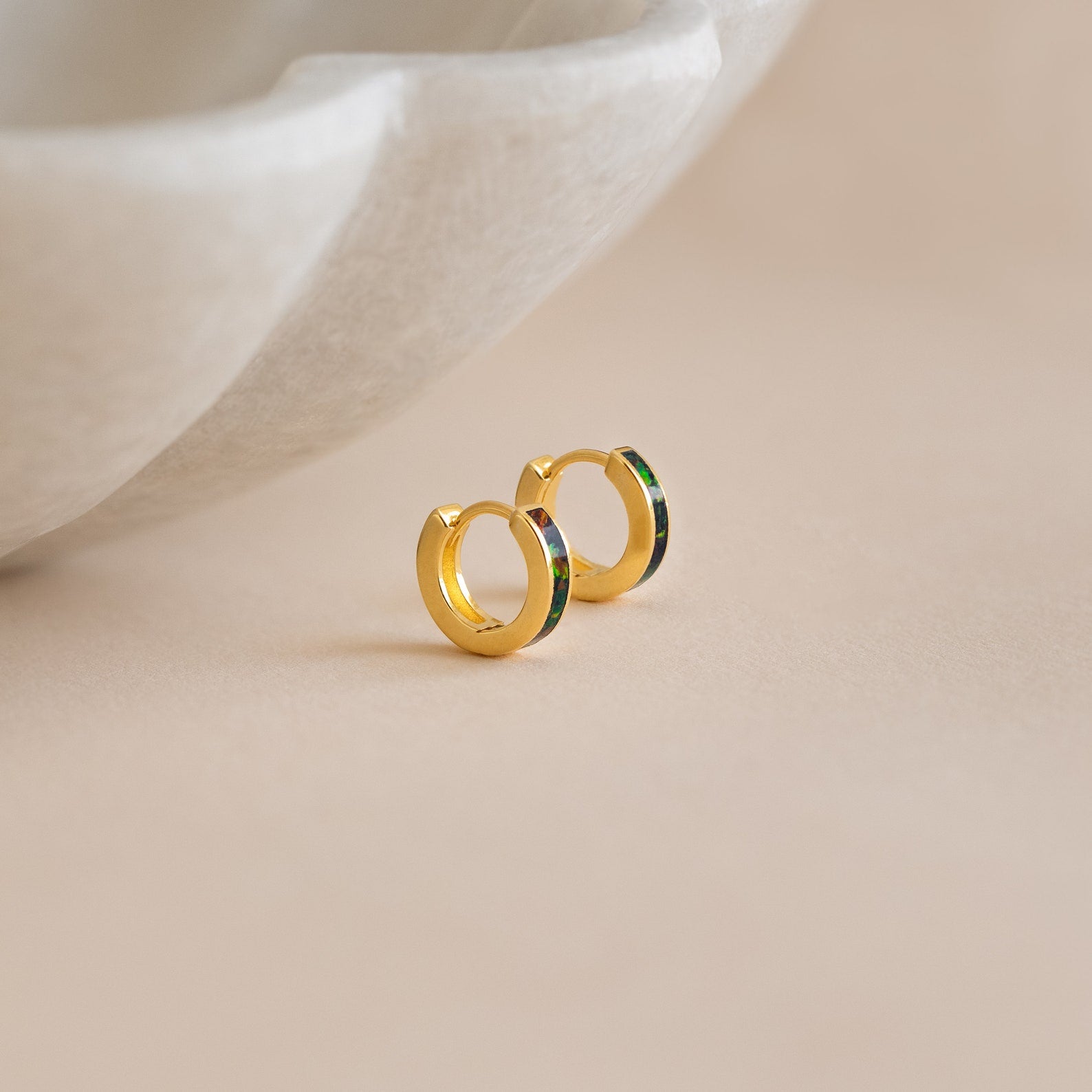 Small gold hoop earrings with a green accent—our Black Opal Inlay Huggies—are displayed on a light beige surface near a marble dish.