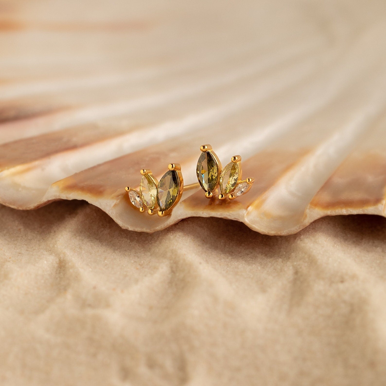 Seagrass Peridot Marquise Studs in gold and green rest on a seashell, with sand visible in the background.
