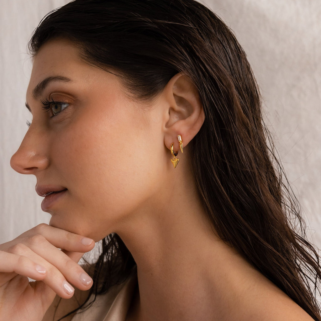 A woman with wet, dark hair wears Shark Tooth Huggies—gold huggie hoop earrings—posing thoughtfully with her hand near her chin for an effortless chic beach jewelry statement.