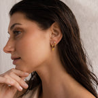 A woman with wet, dark hair wears Shark Tooth Huggies—gold huggie hoop earrings—posing thoughtfully with her hand near her chin for an effortless chic beach jewelry statement.