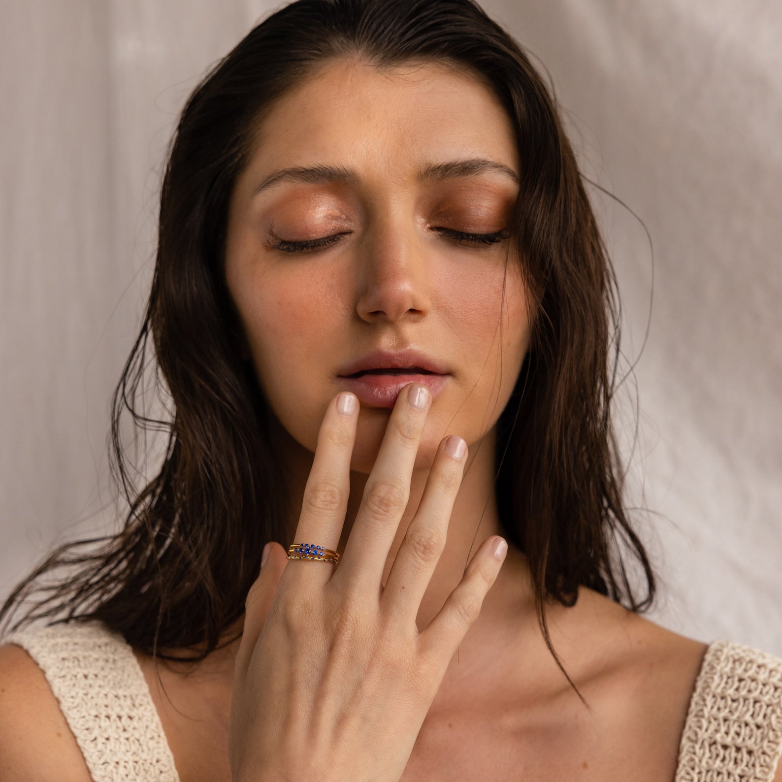A woman with closed eyes touches her lips, wearing a knitted top and the Sapphire Stacking Ring Set adorned with blue sapphires, set against a neutral background.