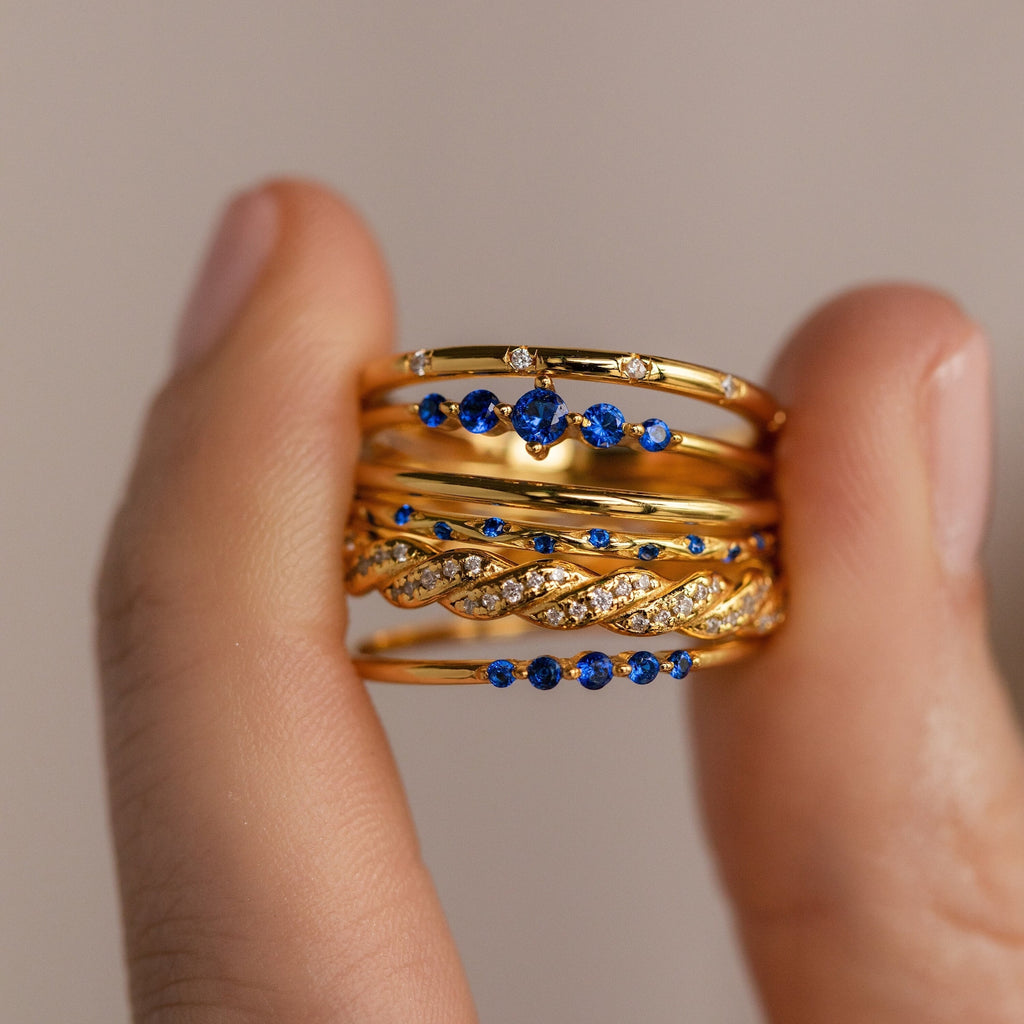 A hand displays the Sapphire Stacking Ring Set—stacked gold rings adorned with blue sapphires and white gemstones for an elegant look.
