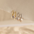 Two pairs of Bridget Pearl Hoops—each with delicate pearl charms, one pair with gold huggies and one with silver—are displayed on a textured surface.