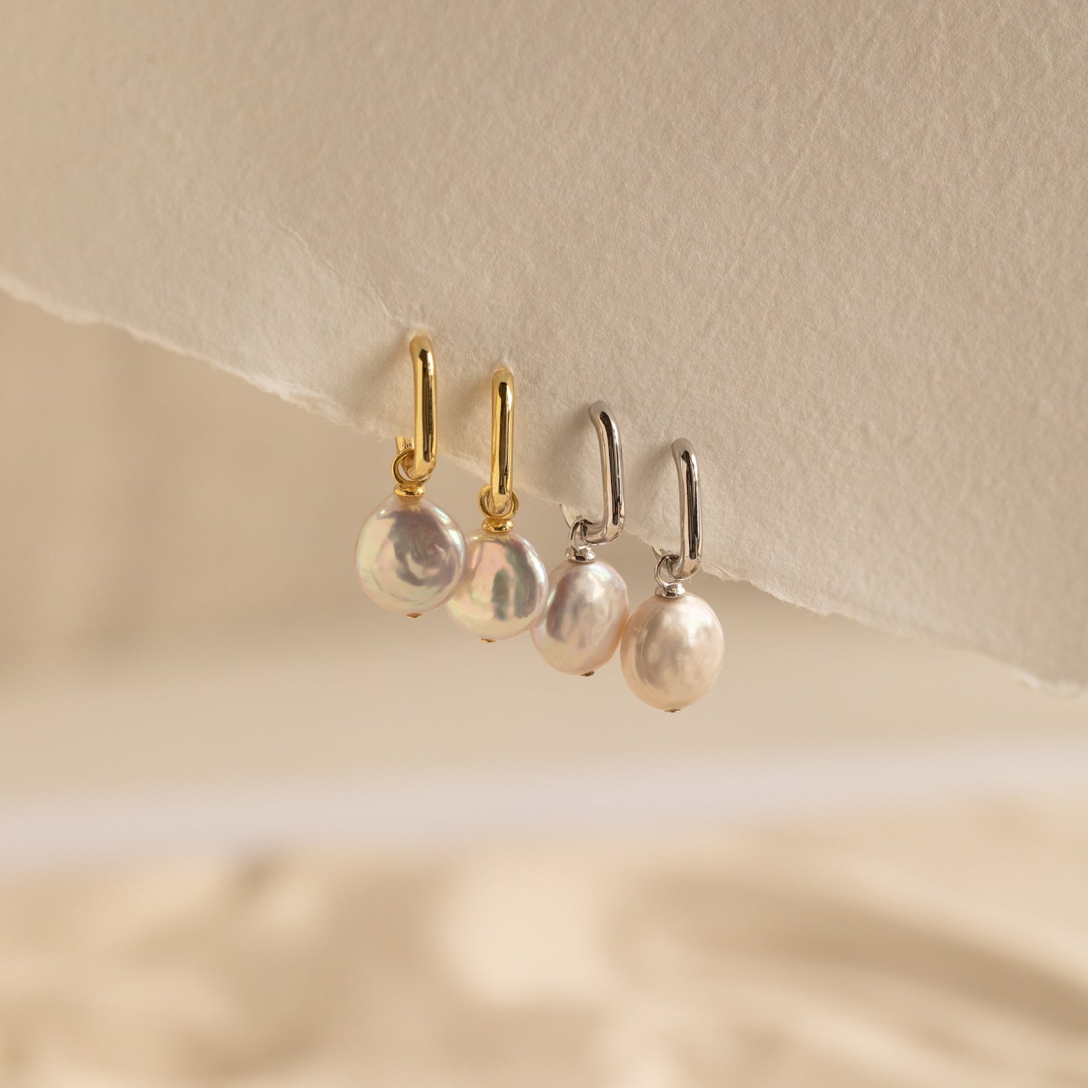 Two pairs of Bridget Pearl Hoops—each with delicate pearl charms, one pair with gold huggies and one with silver—are displayed on a textured surface.