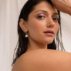 A woman with wet hair, shimmery purple eyeshadow, and a pearl earring poses before a white fabric backdrop, highlighting the elegant Bridget Pearl Hoops.