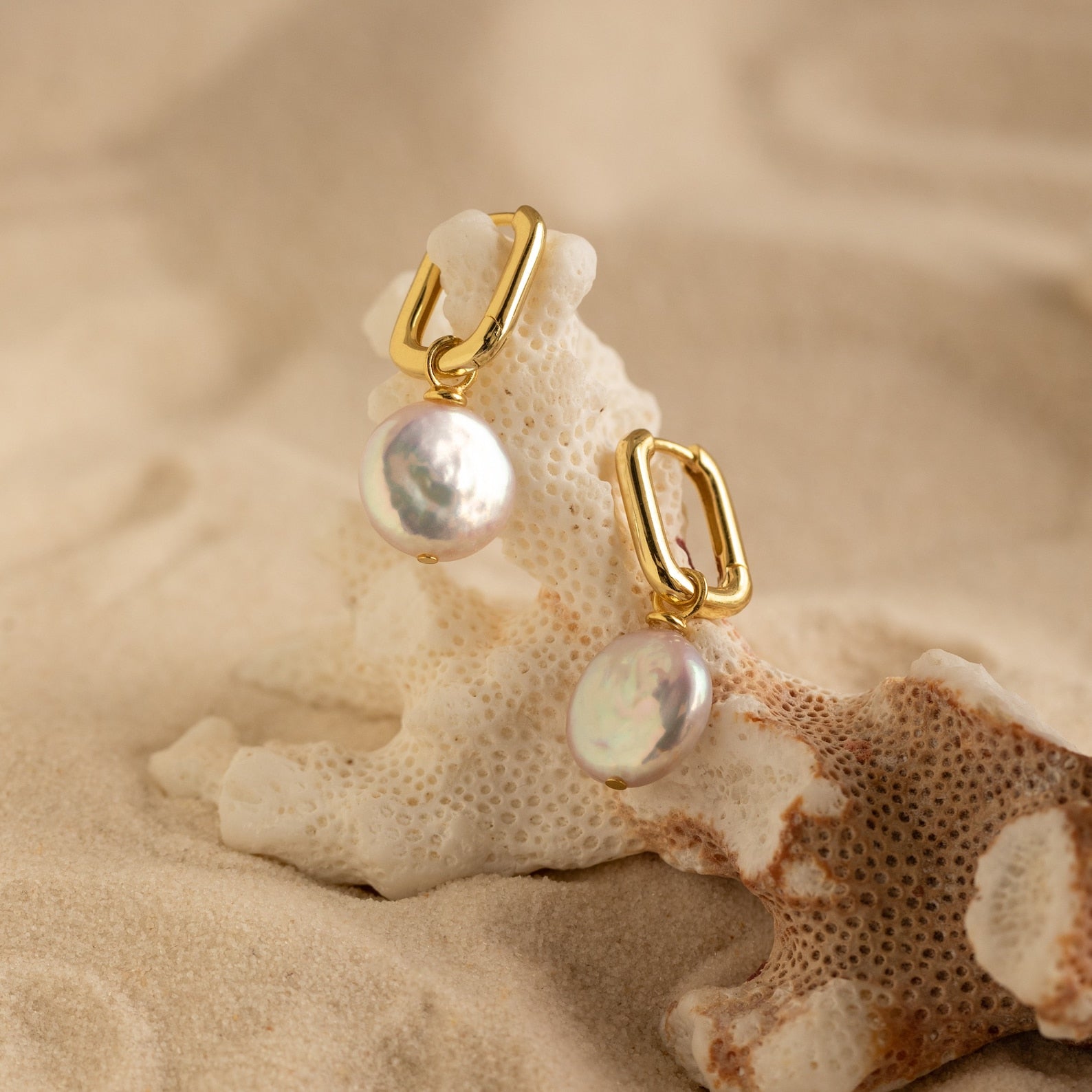 Bridget Pearl Hoops showcase elegant pearl charms on delicate huggie hoops, artfully displayed atop coral and surrounded by sand.