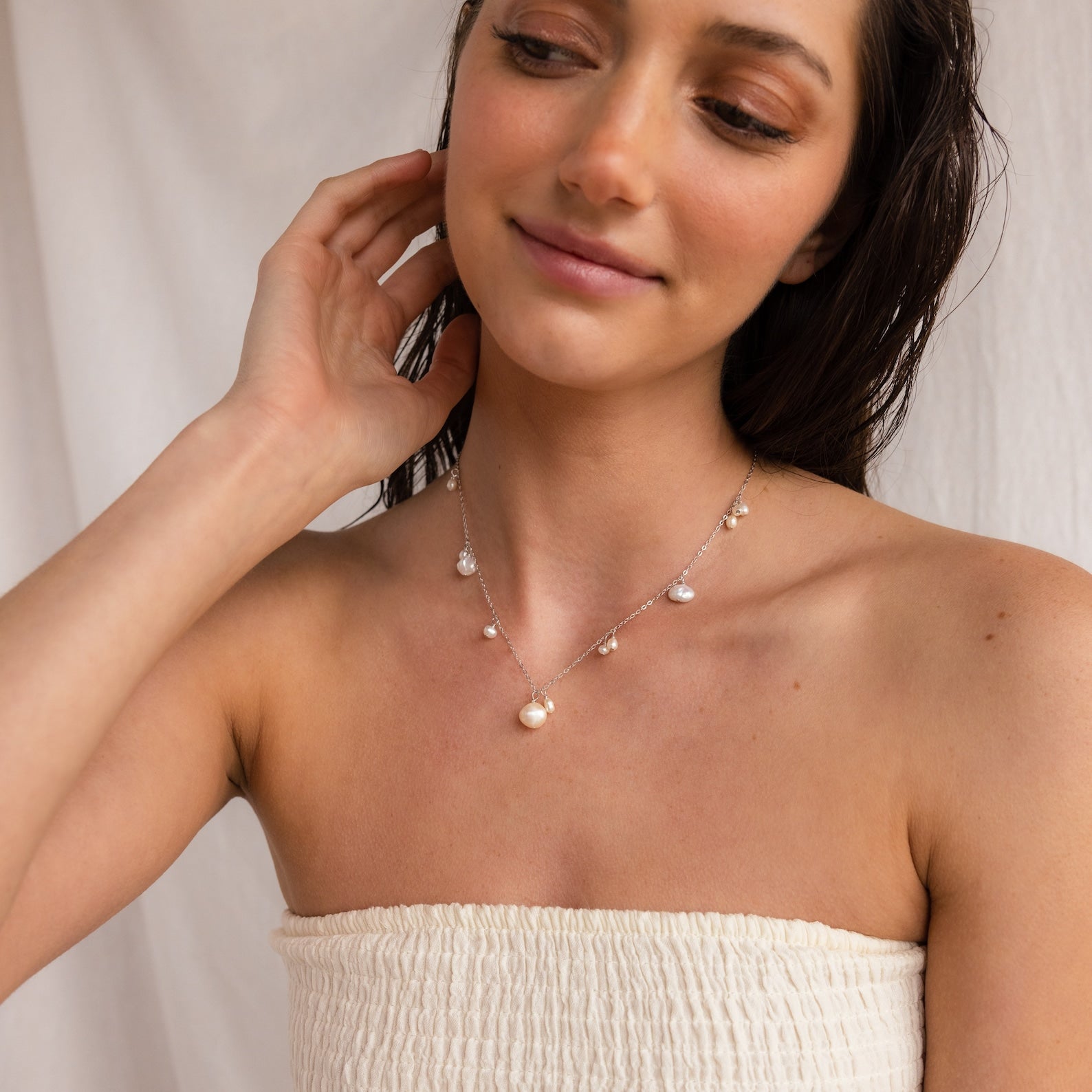 A woman in a strapless top touches her neck, wearing the Baroque Pearl Station Necklace—a minimalist piece with pearl-like charms, ideal for weddings or elegant occasions.