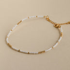 The Dainty Pearl Beaded Anklet features small white and gold beads with a gold chain and clasp, displayed on a beige background.
