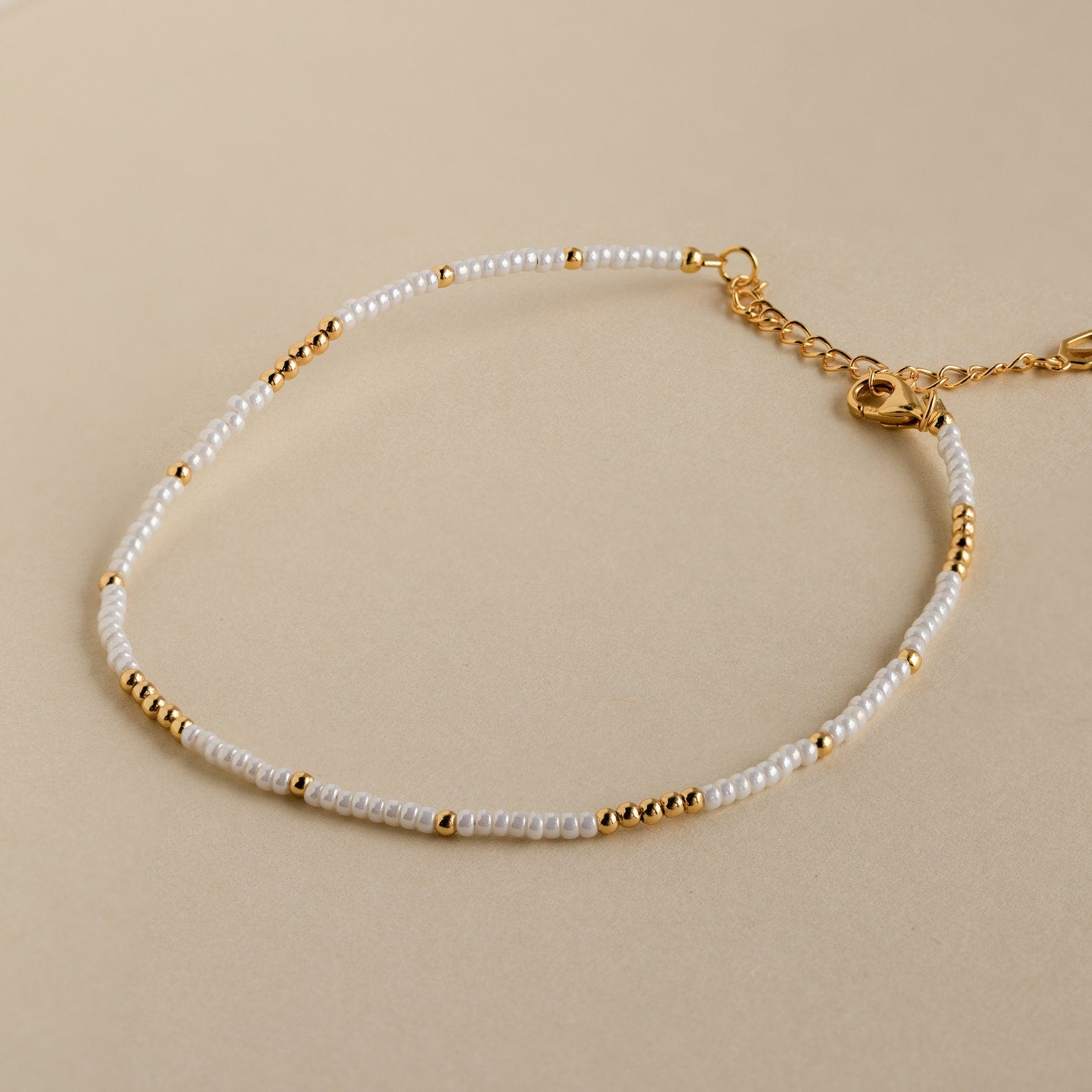 The Dainty Pearl Beaded Anklet features small white and gold beads with a gold chain and clasp, displayed on a beige background.