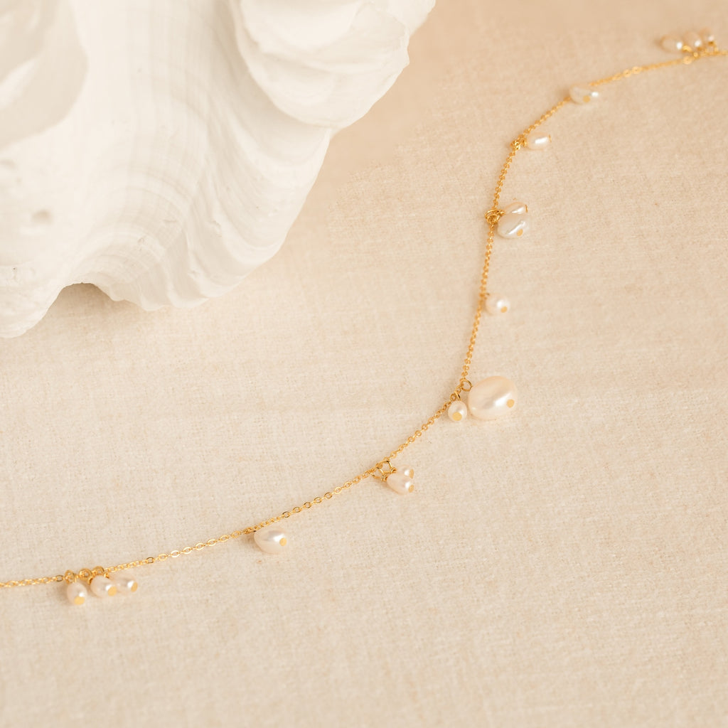 The Baroque Pearl Station Necklace features a delicate gold chain with small pearls, displayed on a cream textured surface near a white shell—an elegant choice for a wedding necklace.
