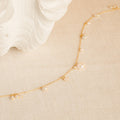 The Baroque Pearl Station Necklace features a delicate gold chain with small pearls, displayed on a cream textured surface near a white shell—an elegant choice for a wedding necklace.