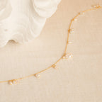 The Baroque Pearl Station Necklace features a delicate gold chain with small pearls, displayed on a cream textured surface near a white shell—an elegant choice for a wedding necklace.