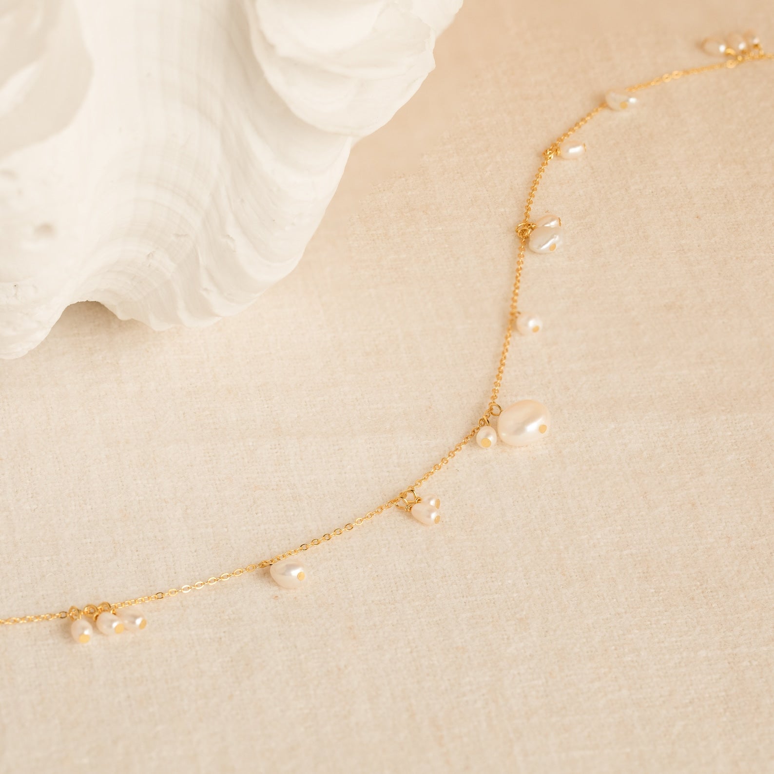 The Baroque Pearl Station Necklace features a delicate gold chain with small pearls, displayed on a cream textured surface near a white shell—an elegant choice for a wedding necklace.