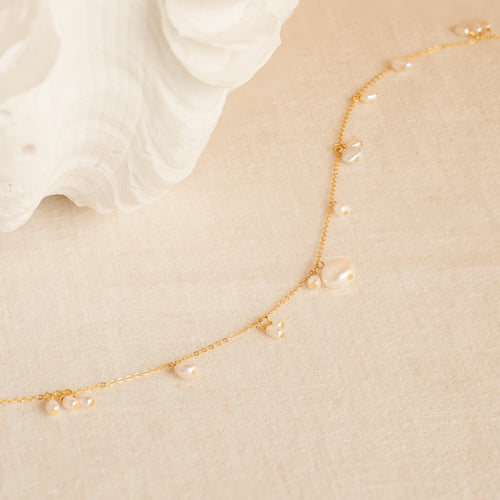 The Baroque Pearl Station Necklace features a delicate gold chain with small pearls, displayed on a cream textured surface near a white shell—an elegant choice for a wedding necklace.