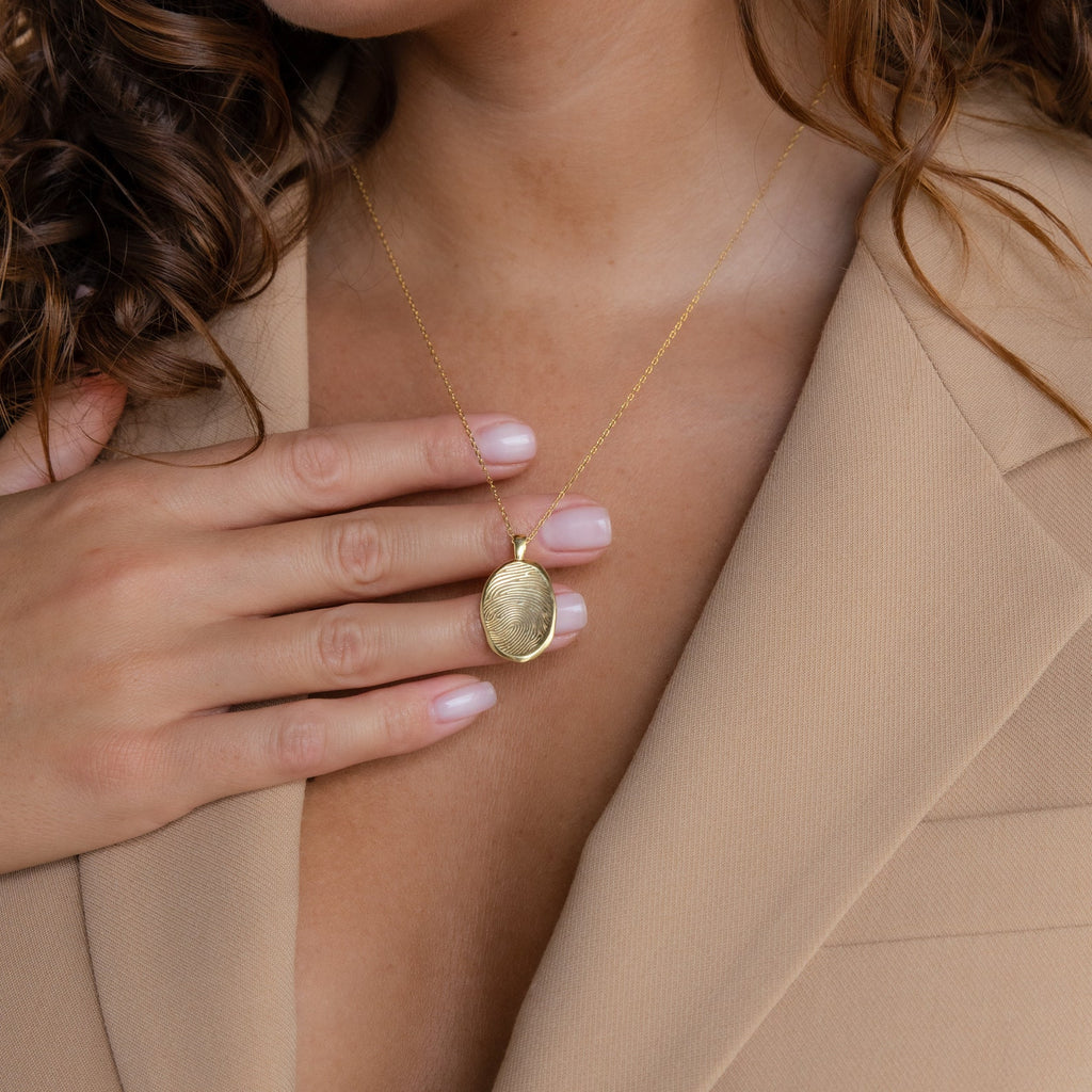 A woman in a beige blazer touches a Custom Fingerprint Necklace in Rose Gold on her chest.