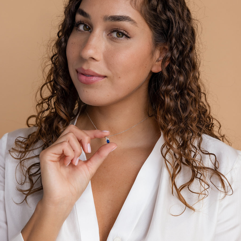 Woman with curly hair in a white blouse holds the Lorelei Sapphire Teardrop Necklace, which features a small blue pendant, against a neutral background.