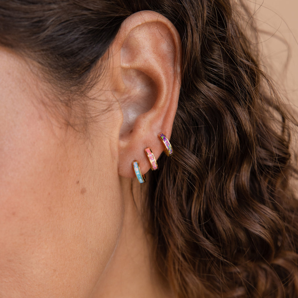 Close-up of a woman’s ear with three small, colorful hoop earrings and curly brown hair, featuring Pastel Opal Inlay Huggies that add a delicate touch of pastel opal jewelry.