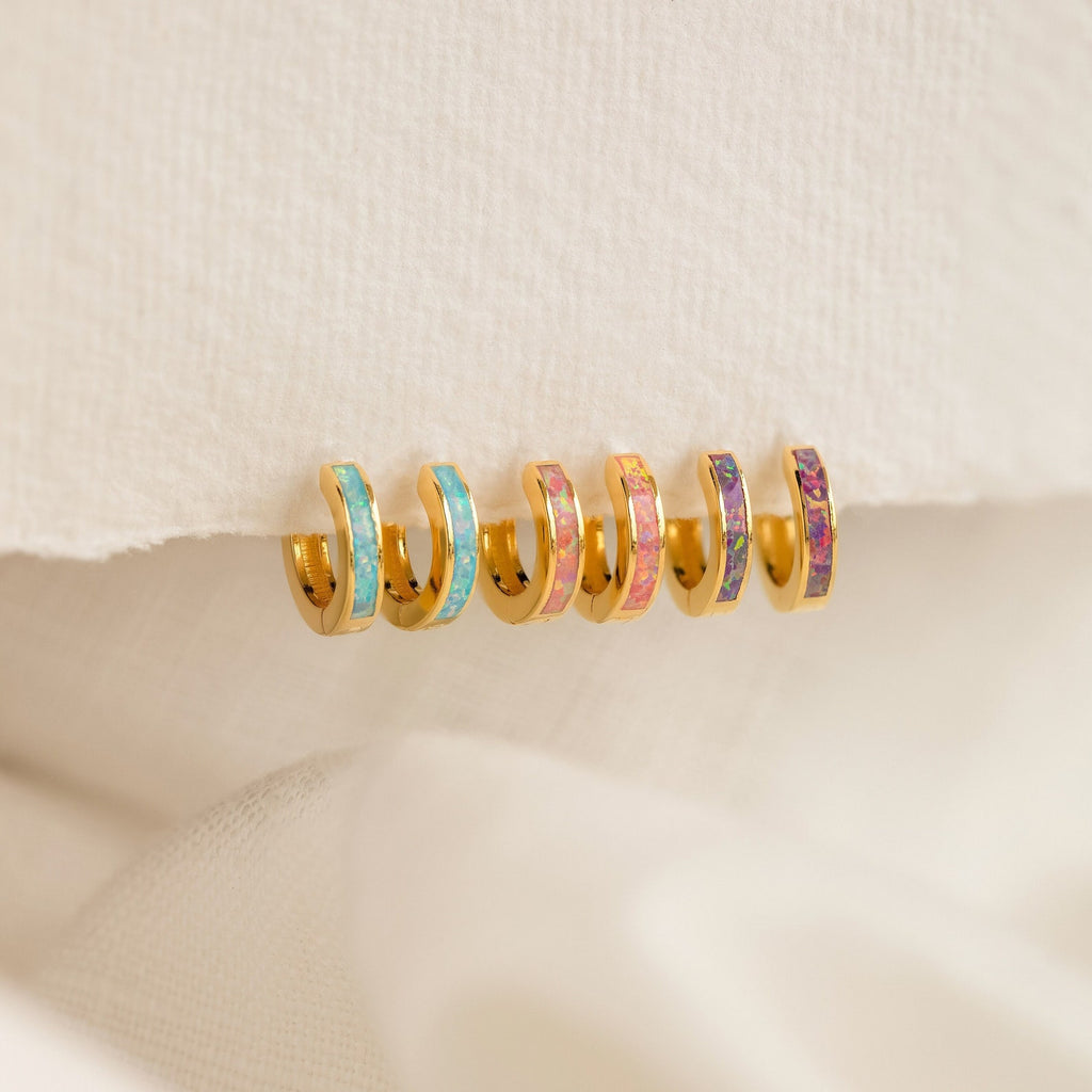 Six Pastel Opal Inlay Huggies—small gold hoops accented with colorful enamel—are displayed on a textured cream fabric background.