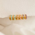 Six Pastel Opal Inlay Huggies—small gold hoops accented with colorful enamel—are displayed on a textured cream fabric background.