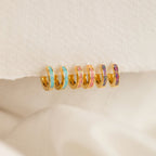 Six Pastel Opal Inlay Huggies—small gold hoops accented with colorful enamel—are displayed on a textured cream fabric background.