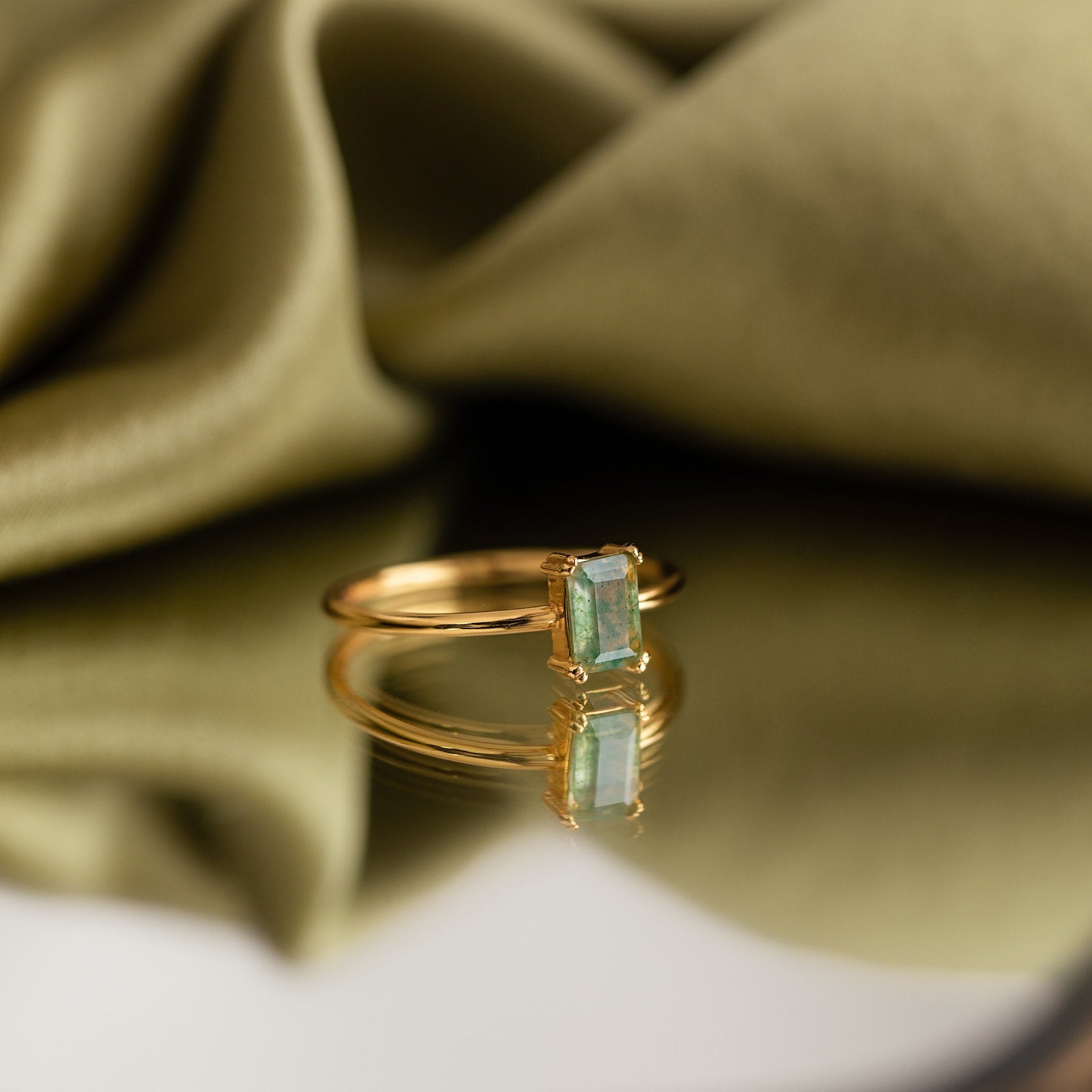 The Agate Emerald Ring placed on a reflective surface with olive green fabric draped behind, creating a luxurious look.