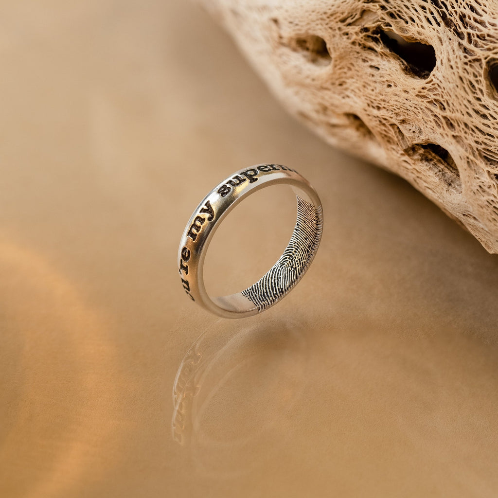 The Half Round Fingerprint Ring, personalized with engraved text and a fingerprint, sits on a smooth beige surface near textured stone, highlighting the unique charm of custom jewelry.