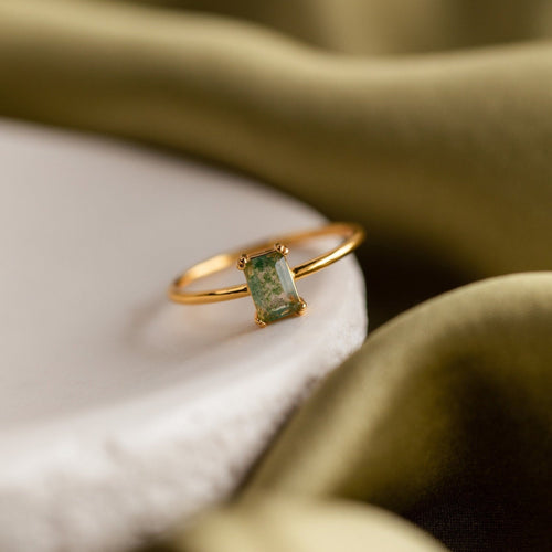 A close-up photograph of the Agate Emerald Ring resting elegantly on a smooth white surface with olive green fabric draped in the background. The ring features a slender, polished gold band and a rectangular-cut agate stone with natural emerald green and earthy moss-like inclusions, held by four delicate prongs