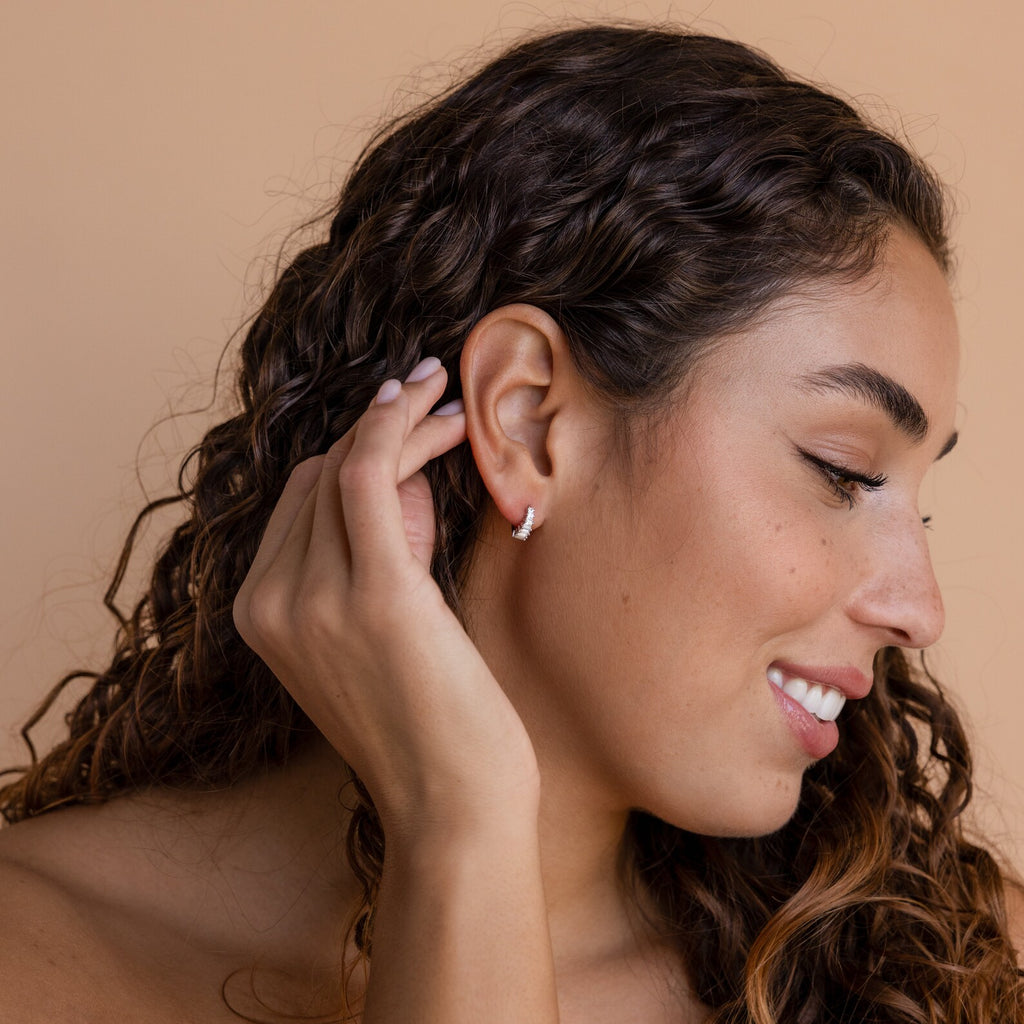 Model with long curly hair wearing a single silver Art Deco Huggie earring, smiling with her hand near her face.