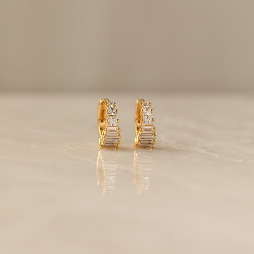 Close-up of a pair of gold Art Deco Huggie earrings on a marble surface, featuring a symmetrical arrangement of baguette and round-cut diamonds for a tiered, structured look.