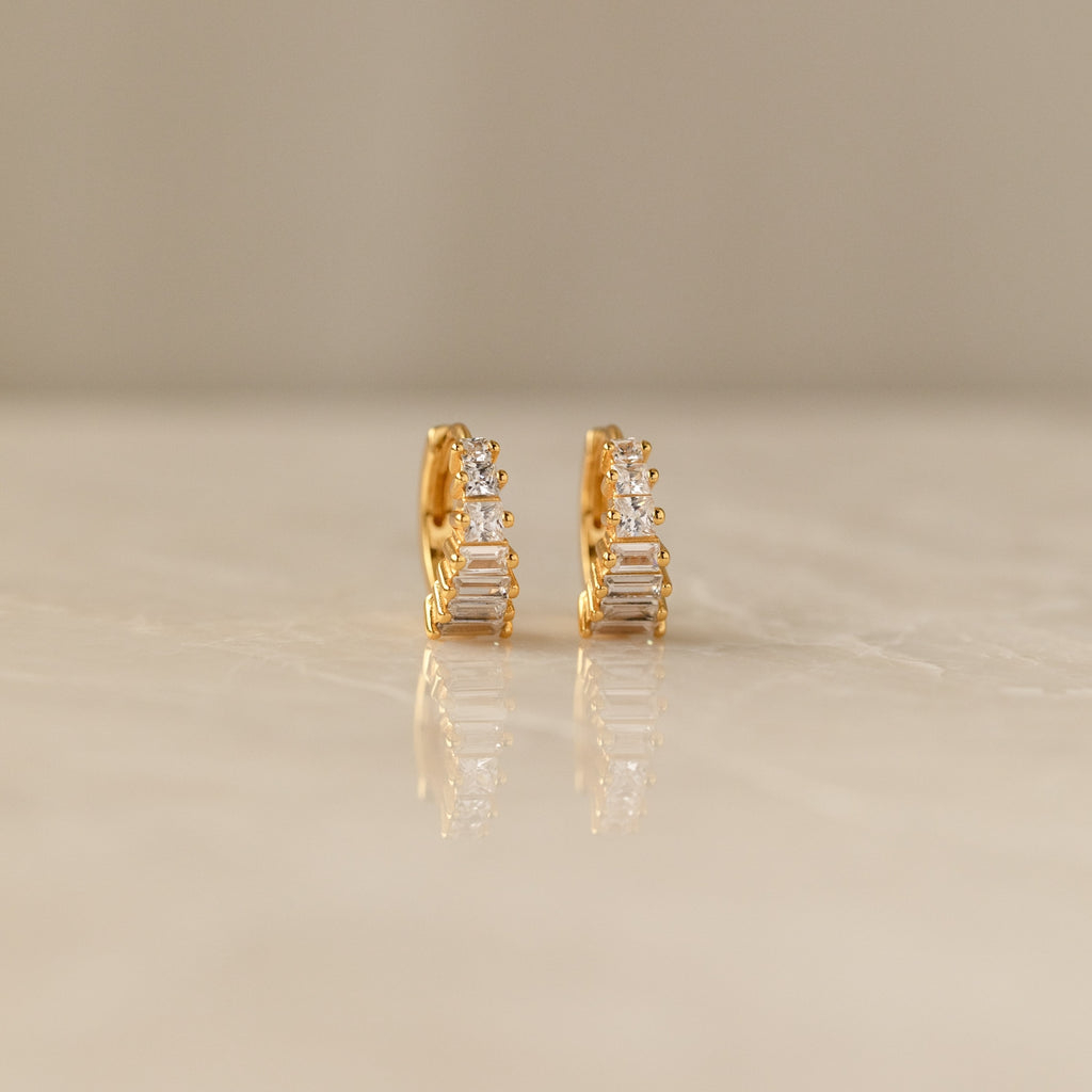 The Art Deco Huggies & Ring Set features gold hoops with vertical rectangular and round clear stones, evoking classic Art Deco style, elegantly displayed on a smooth neutral surface.