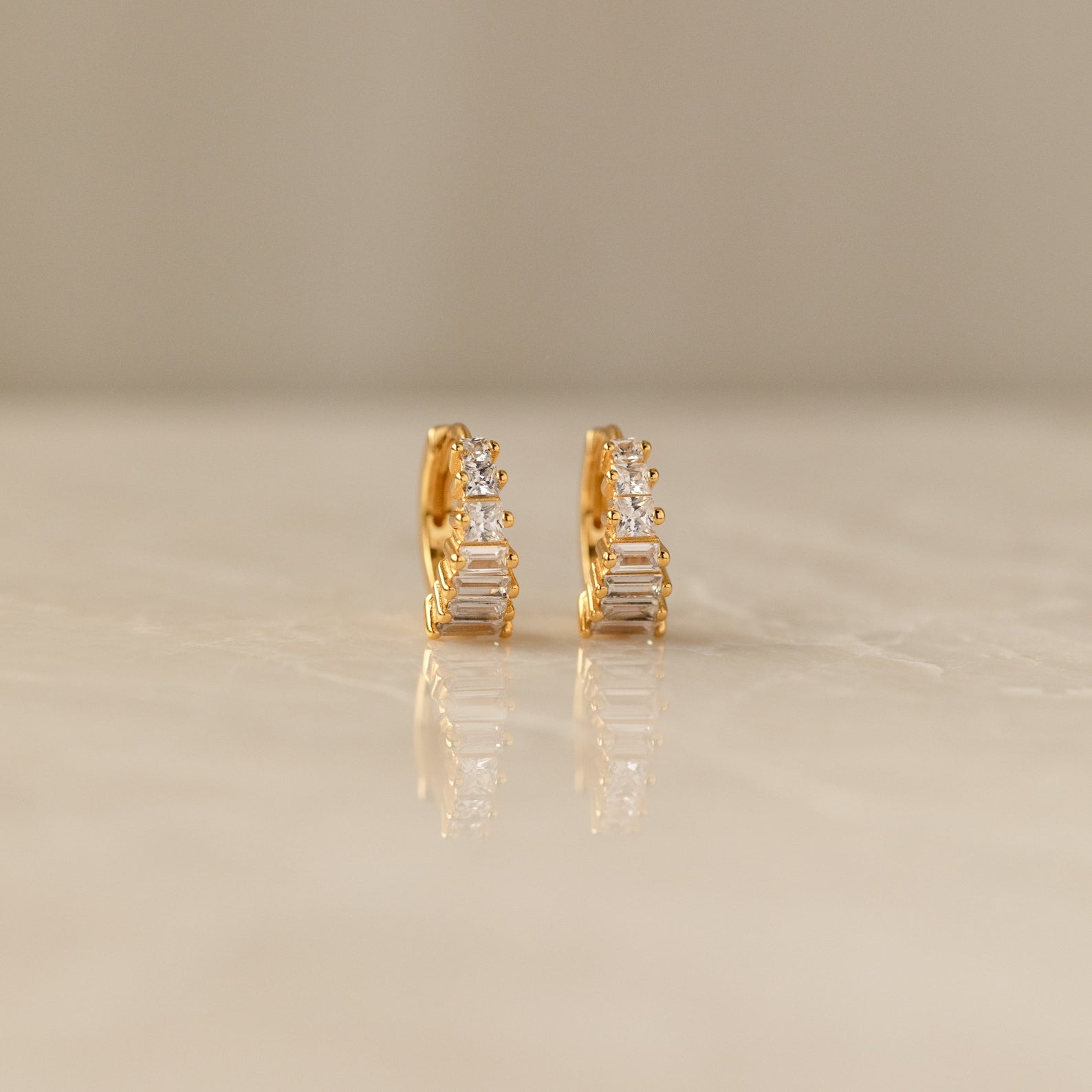 The Art Deco Huggies & Ring Set features gold hoops with vertical rectangular and round clear stones, evoking classic Art Deco style, elegantly displayed on a smooth neutral surface.