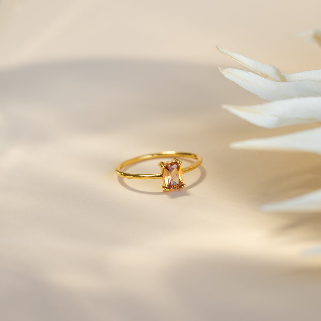 The Citrine Emerald Ring is displayed on a soft beige surface beside white dried foliage, showcasing its elegant emerald-cut gemstone.