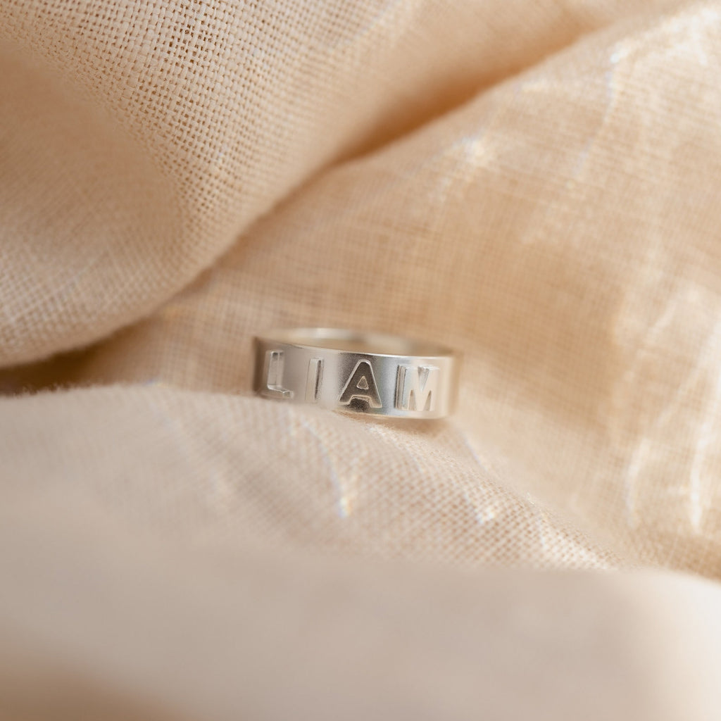 3D Name Band sterling silver ring with bold 3D engraved letters spelling LIAM on textured fabric background
