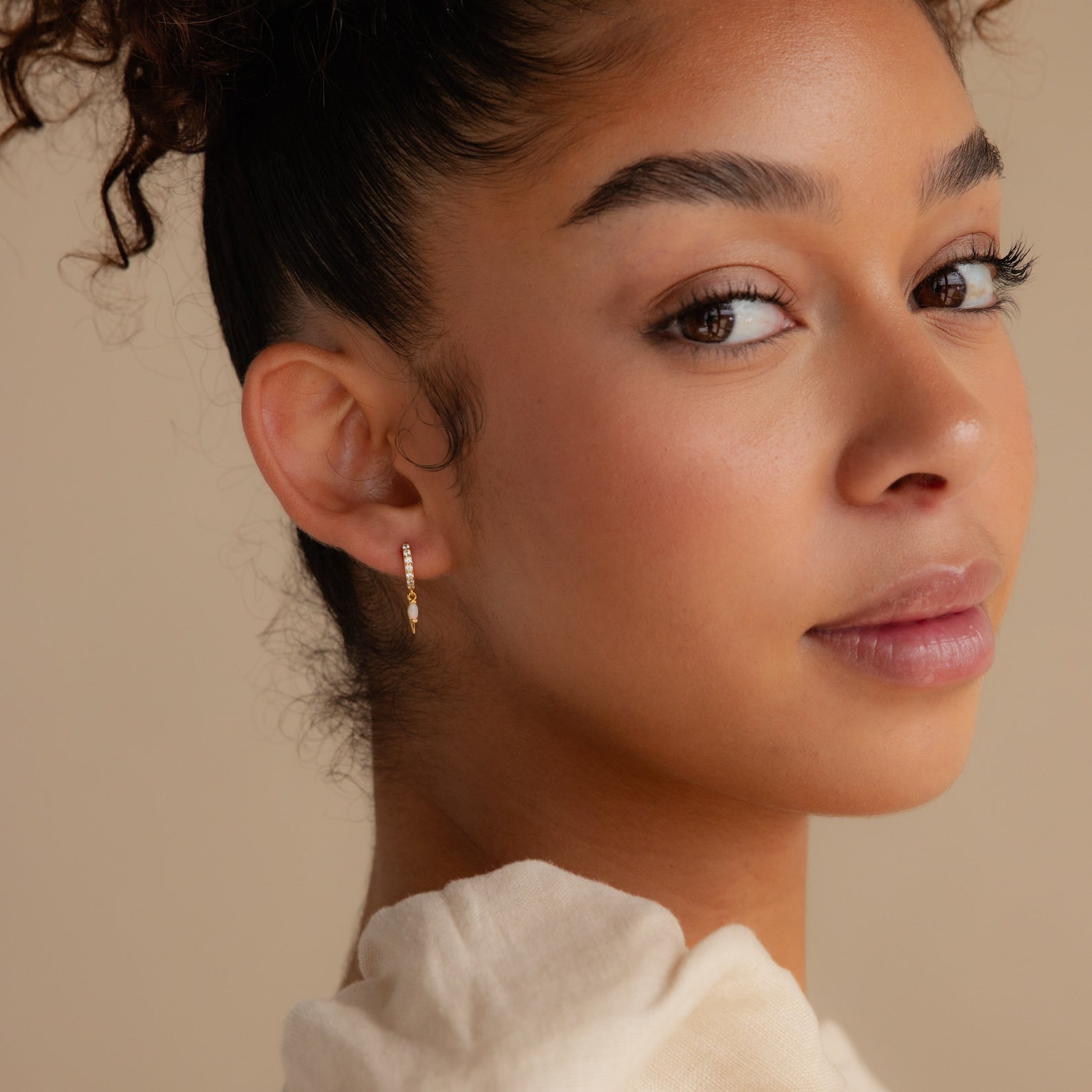 A woman with curly hair looks over her shoulder, wearing Raine Opal Dagger Huggies on a neutral background.