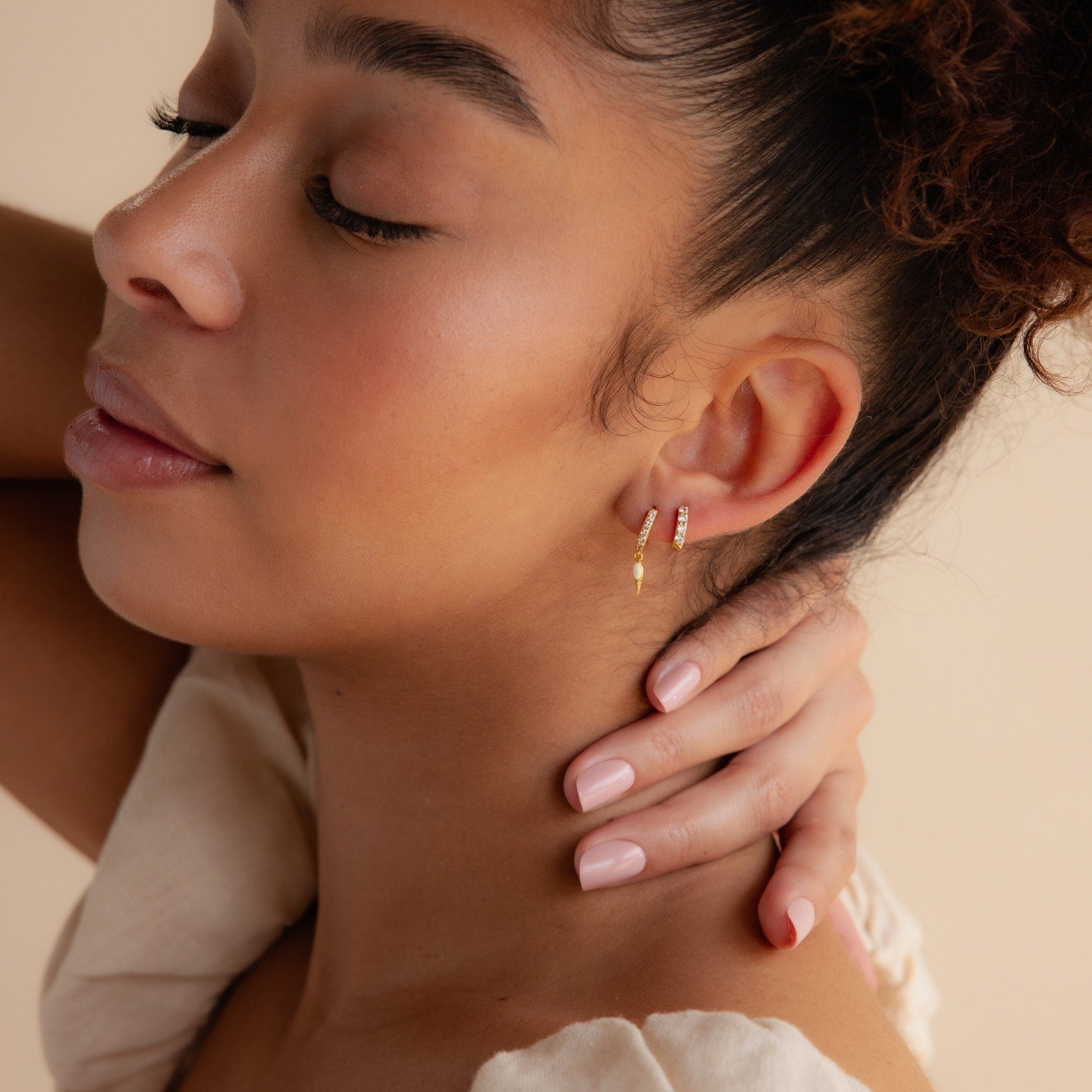Woman with closed eyes touches her neck, wearing the Raine Opal Dagger Huggies and a cream-colored top, effortlessly embodying their bohemian elegance.