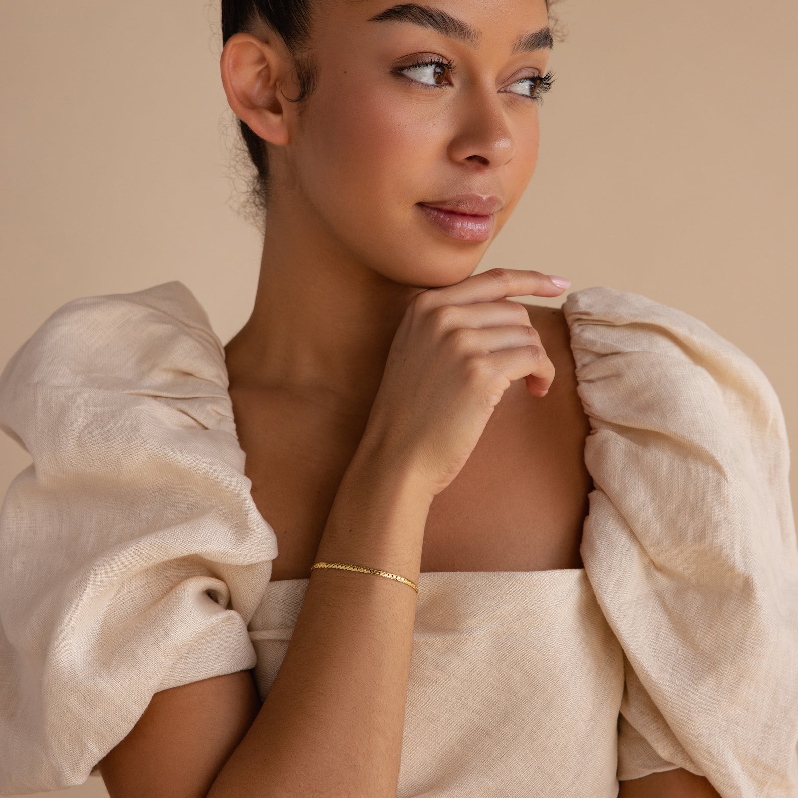 Woman in a cream puff-sleeve dress looks to the side, resting her hand near her chin, styled with a minimalist Snake Chain Bracelet in gold.