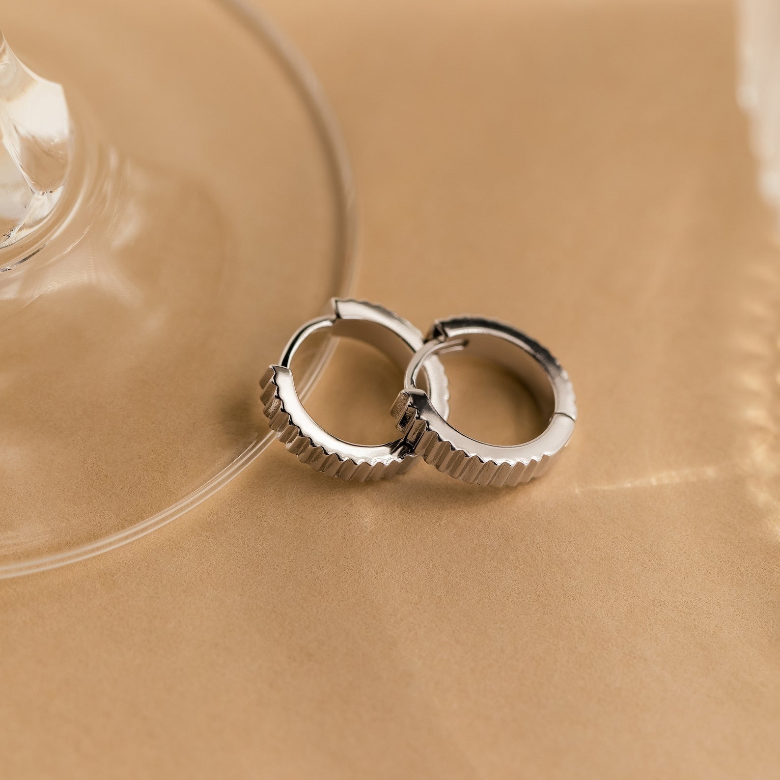 Two silver Ribbed Huggies with ridged edges rest on a beige surface beside a glass object—ideal for anyone who loves dainty earrings with subtle texture.