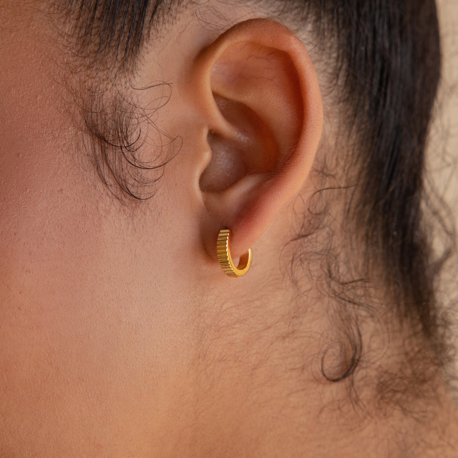 Close-up of a person's ear wearing Ribbed Huggies—small, ribbed hoop earrings perfect for those who love dainty earrings with subtle texture.