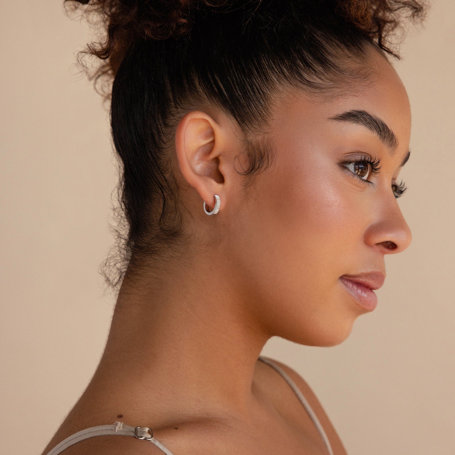 Woman with curly hair in a bun, wearing dainty Ribbed Huggies, looks to the right against a beige background.