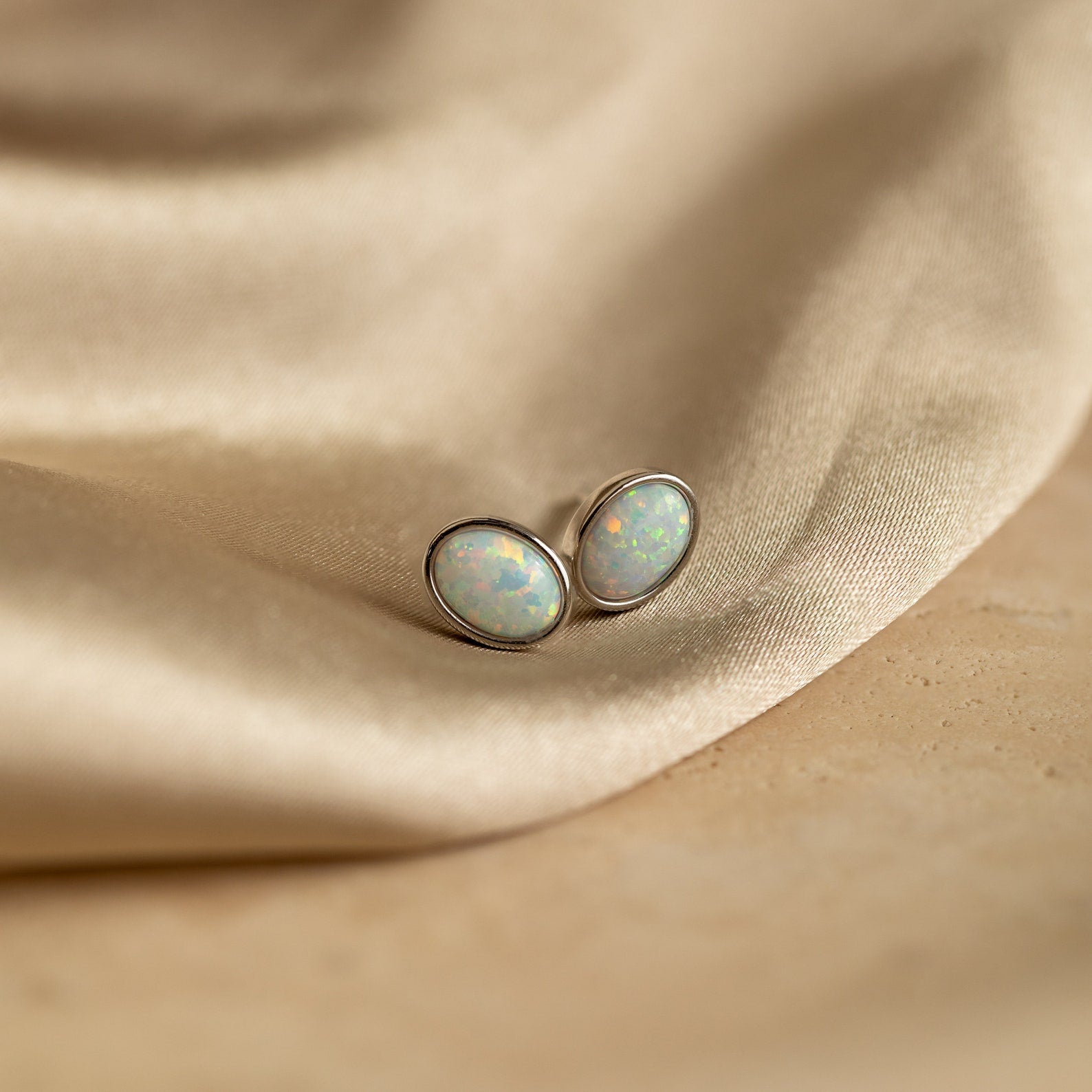 Oval Opal Studs featuring a luminous white opal stone set in silver trim, beautifully displayed on light beige satin fabric.