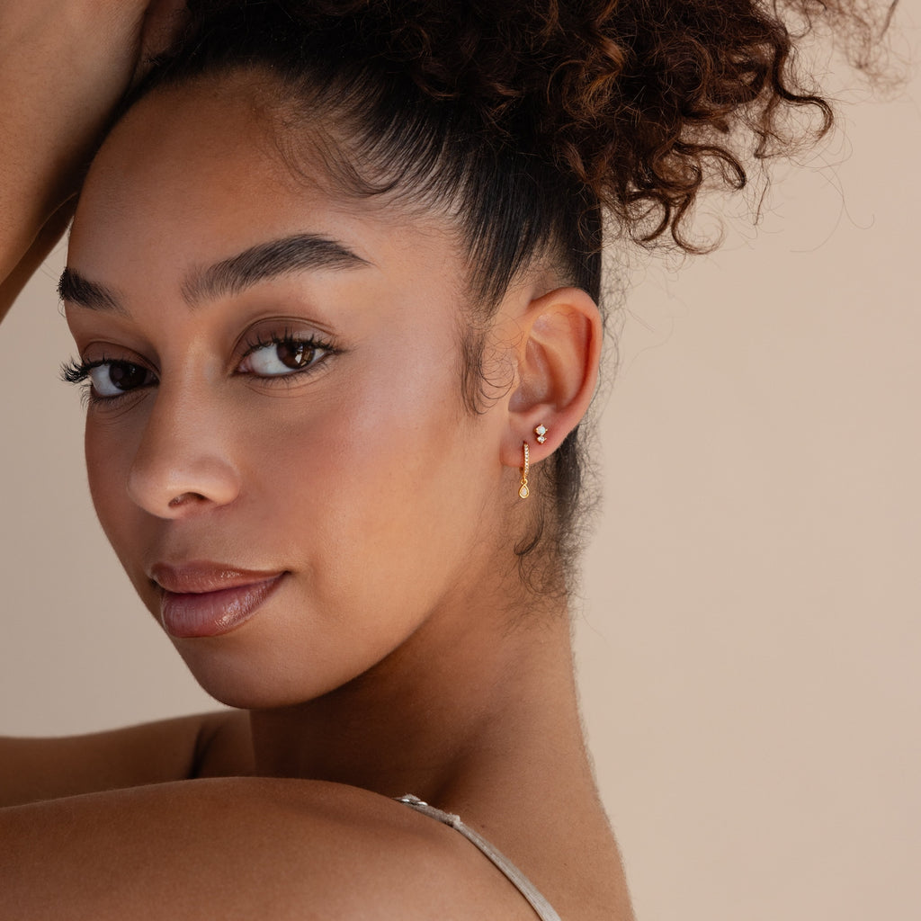 A woman with curly hair and gold earrings poses with her arm raised, showcasing elegant Opal Drop Huggies while looking at the camera against a beige background.