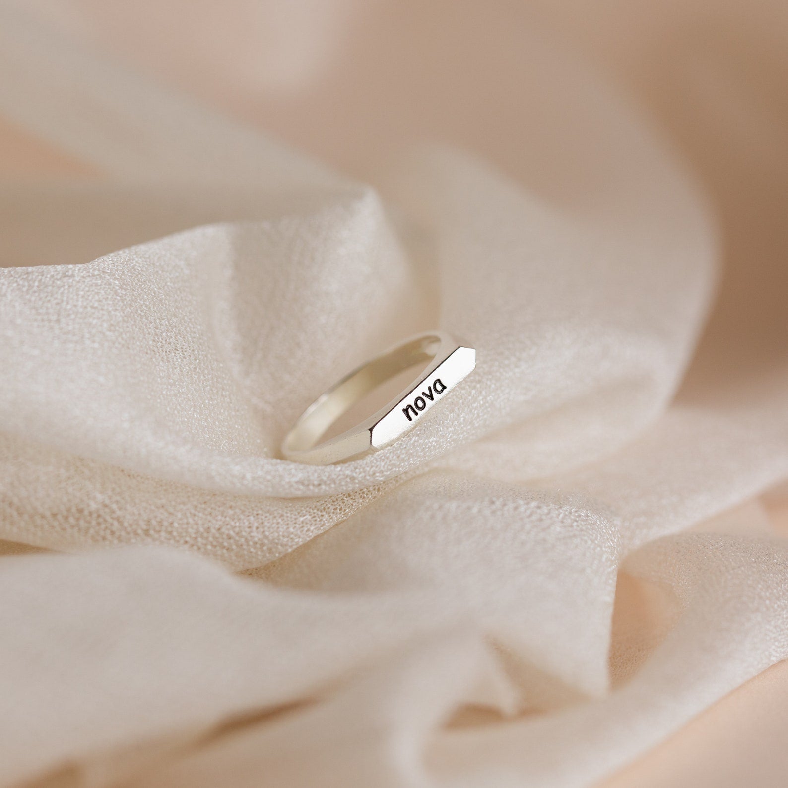 The Thin Name Signet Ring, engraved with "nova," rests on soft beige fabric—perfect for stacking or wearing as a sleek signet bar style.