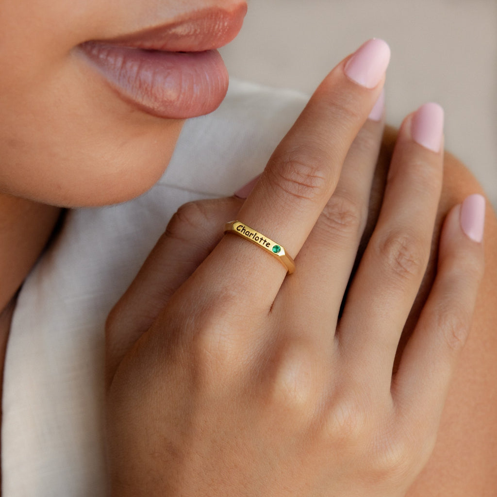 A woman with light pink nails wears the Thin Name Signet Ring engraved with "Charlotte" and a small green gem, perfect for stacking in a classic bar signet style.