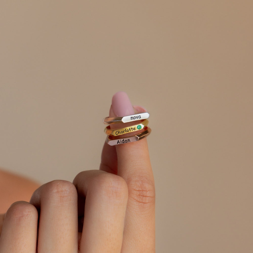 A hand displays three Thin Name Signet Rings—engraved with Nova, Charlotte, and Aidan—stacked on the index finger against a beige background.