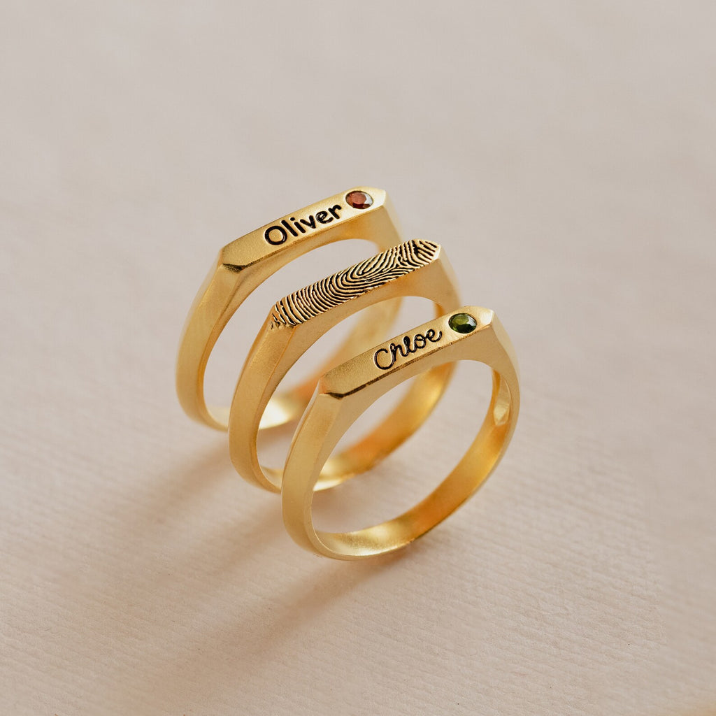 Three Thin Name Signet Rings, each gold, engraved with a name and fingerprint, set with a small gemstone, displayed on a light background.