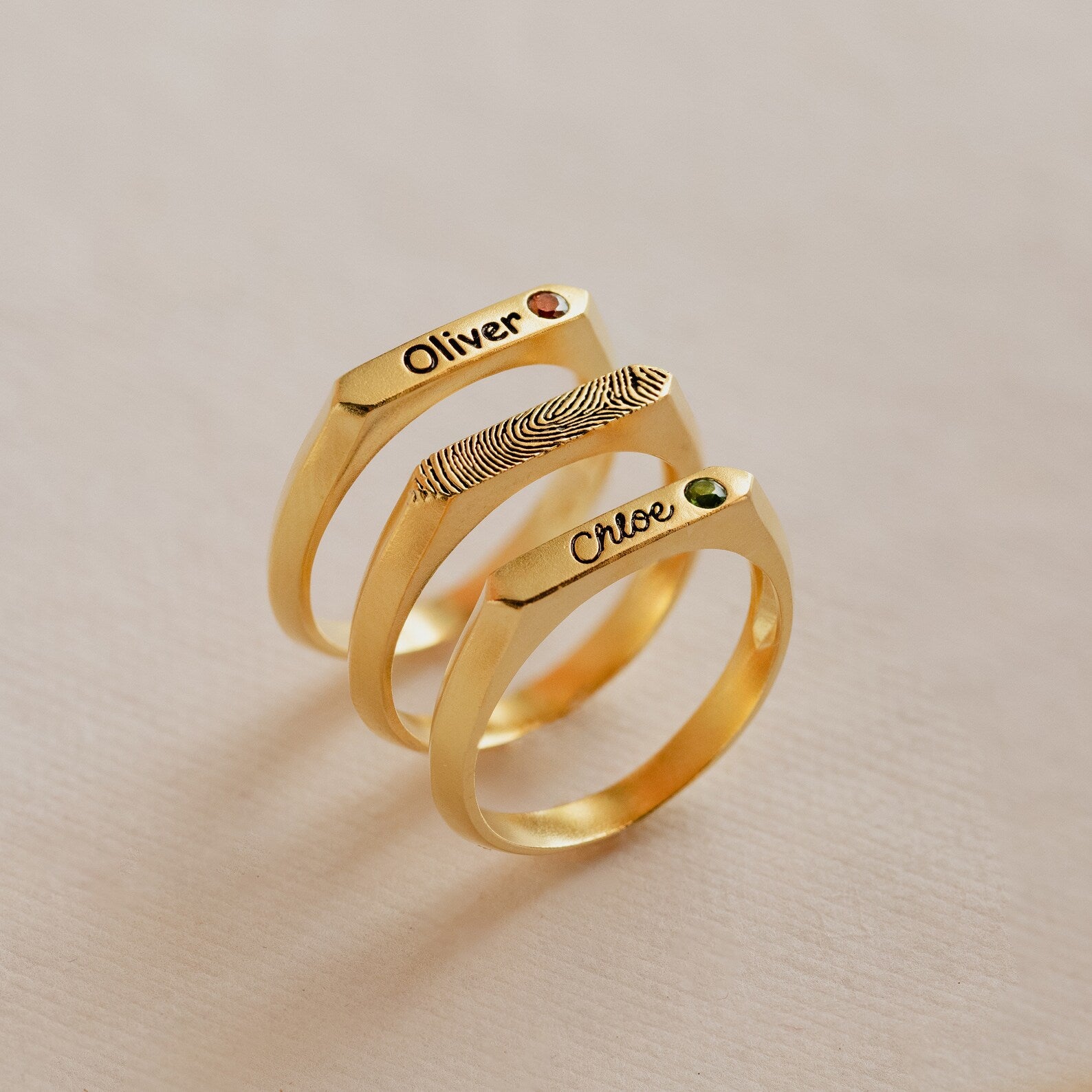 Three Thin Name Signet Rings, each gold, engraved with a name and fingerprint, set with a small gemstone, displayed on a light background.