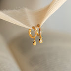 Opal Drop Huggies—gold huggie hoops with delicate opal drops—are softly lit as they hang from a piece of beige fabric.