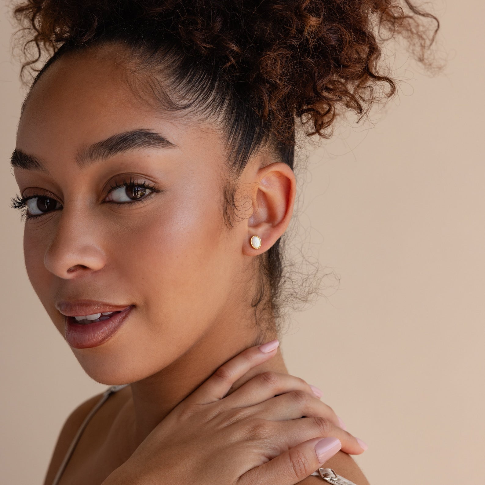 A woman with curly hair smiles warmly, her hand on her shoulder, showcasing elegant Oval Opal Studs against a neutral background.