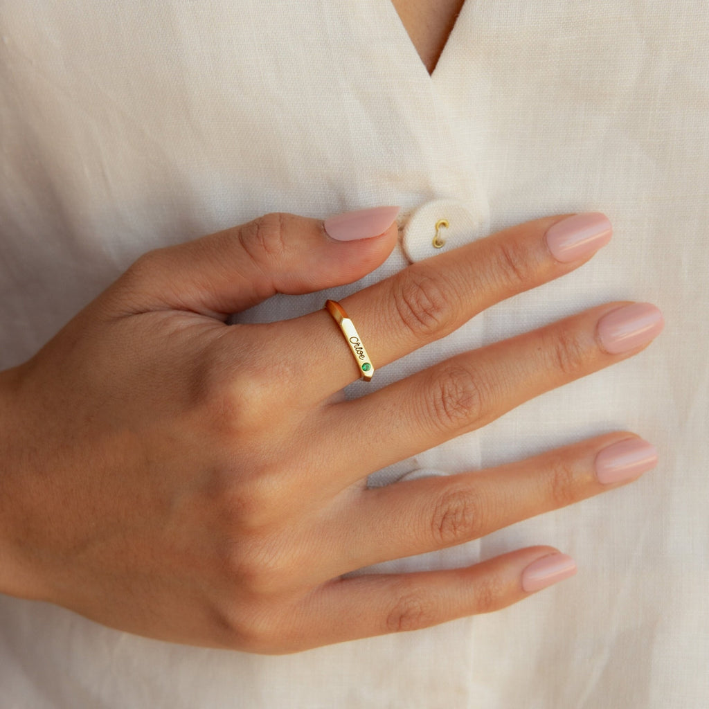 A hand with light pink nails displays the Paris Thin Name Signet Ring in Rose Gold, featuring a sleek design, resting gently on a white shirt.