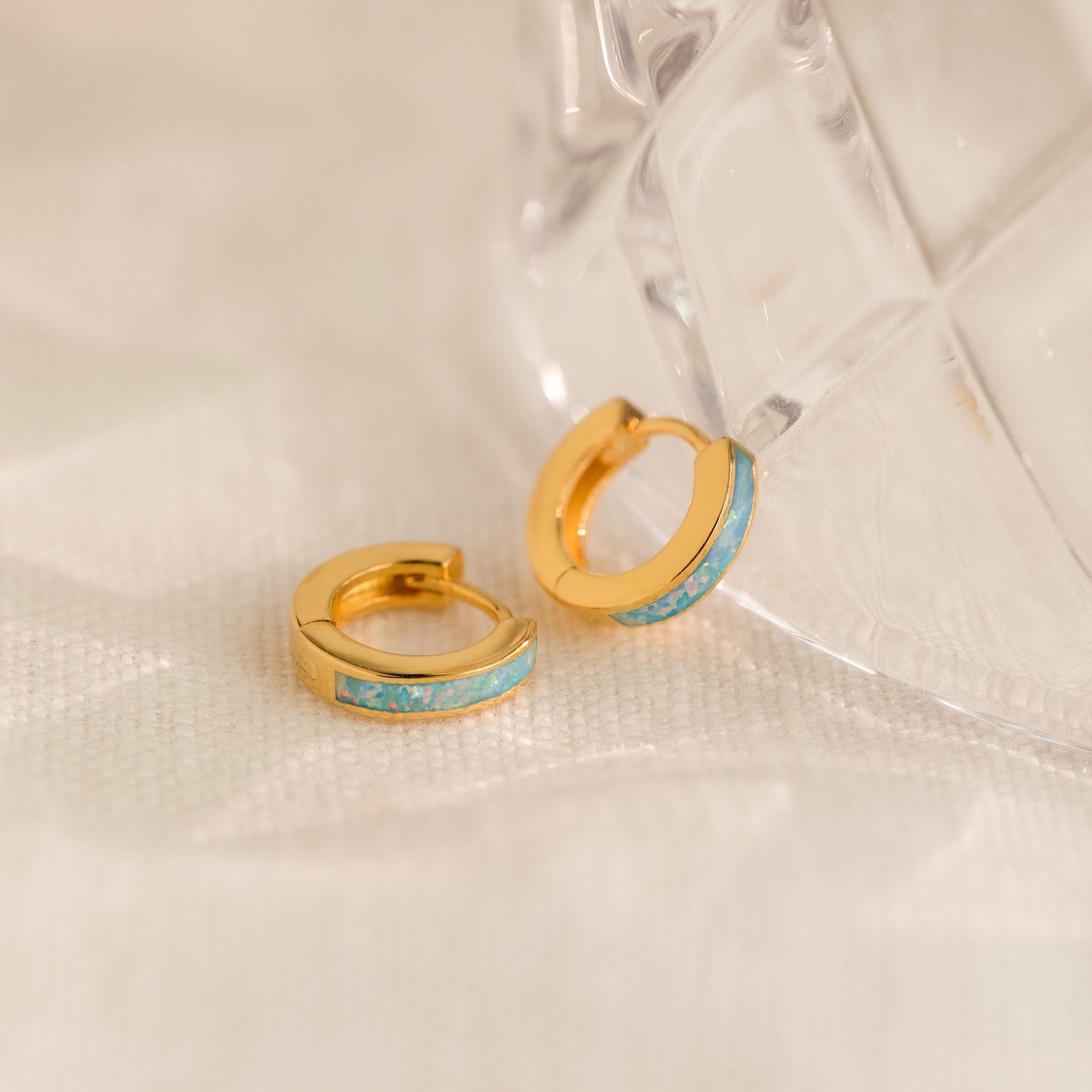 Ocean Opal Inlay Huggies, featuring gold hoops with opal inlay, make elegant small hoop earrings or chic wedding jewelry. Shown beside a clear textured glass on a light fabric surface.